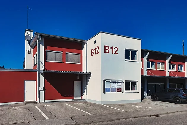 Gebäude des Therapiezentrums B12 in Lechbruck am See: weißer Gebäudeteil mit rotem Schriftzug B12, daneben rote Fassade, davor Parkplätze