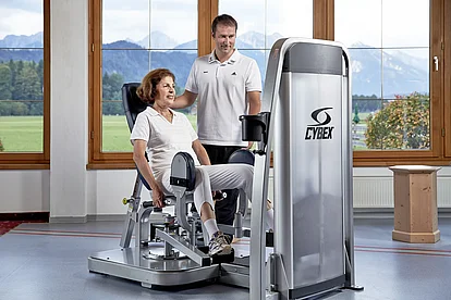 Patientin trainiert an einem Cybex-Kraftgerät, der Therapeut begleitet sie, im Hintergrund Bergblick