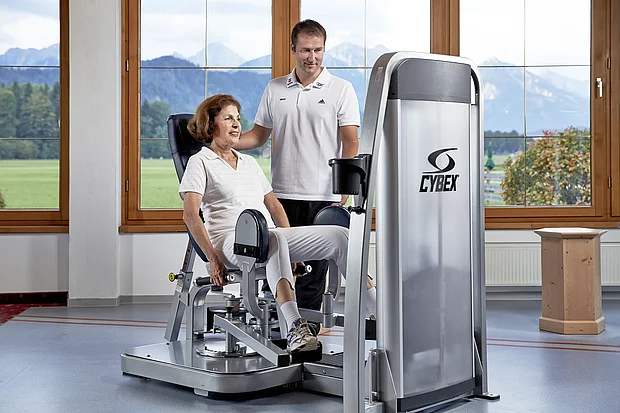 Patientin trainiert an einem Cybex-Kraftgerät, der Therapeut begleitet sie, im Hintergrund Bergblick