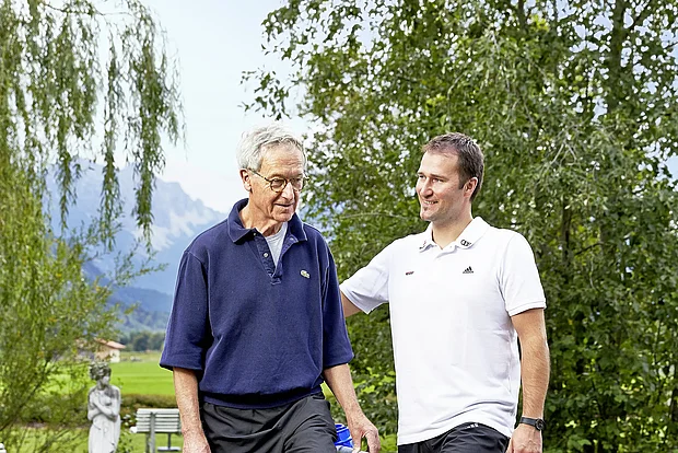 Therapeut im Gespräch mit einem älteren Patienten im Garten der Fachklinik König Ludwig