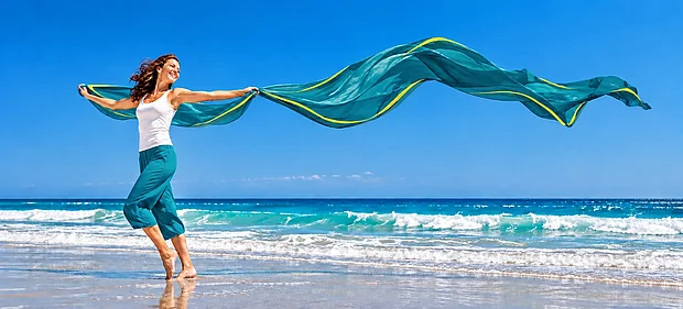Lächelnde Frau läuft barfuß am Strand und hält ein langes türkisfarbenes Tuch, das hinter ihr im Wind weht